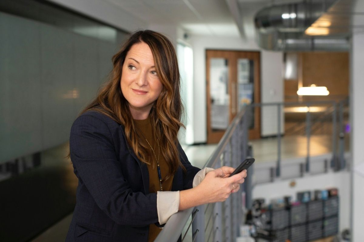 women looking into the distance stood leaning on barrier on her phone